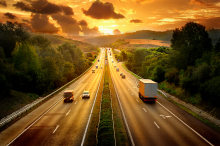 Several cars drive early in the morning at sunset on a four-lane road surrounded by trees.