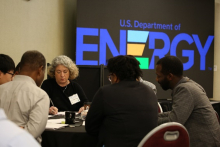 People discussing at a table in front of a DOE logo on a projector screen