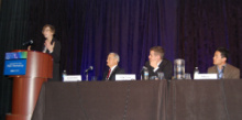 Person standing at a lectern addressing an audience, next to a long table with three more people sitting at it.