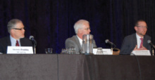 Three men in business attire sitting at a long table addressing an audience.