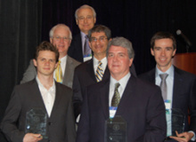 Six men in business attire standing together facing the camera, with award trophies in their hands.