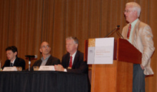 Man in a suit and tie standing at a lectern addressing an audience, next to a long table with three more people sitting at it.