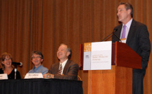 A man standing at a lectern addressing an audience, next to a long table with three more people seated at it.