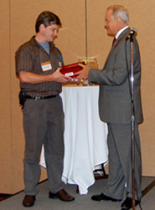 Two men in a business attire standing facing each other, one giving an award to the other.
