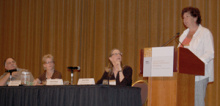 A woman standing at a lectern addressing an audience, next to a long table with three other presenters seated there.