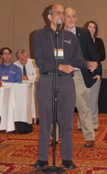 Man standing at a microphone with other people behind him in the background.