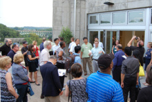 People standing outside a large building, listening to a speaker.