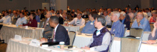 People in business attire sitting at long tables in a convention hall.