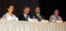 Four people in business attire seated at a long table as presenters in a convention hall.