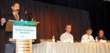 A man standing at a lectern addressing an audience, next to a long table with three other people seated at it.