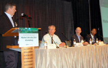 A man standing at a lectern addressing an audience, next to a long table with three other people seated at it.