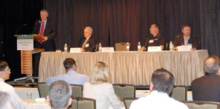 A man standing at a lectern addressing an audience, next to a long table with three other people seated at it.