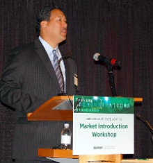 Man in a suit and tie standing at a lectern addressing an audience with a sign on the lectern saying "Market Introduction Workshop."
