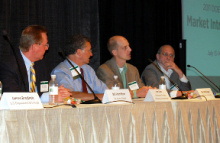 Four men sitting at a table in front of an audience, with microphones, addressing them.