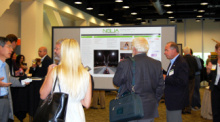 People standing in a convention hall, their backs to the camera, and a large display screen in the background.