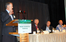 A man standing at a lectern addressing an audience, next to a long table with three other men seated at it.