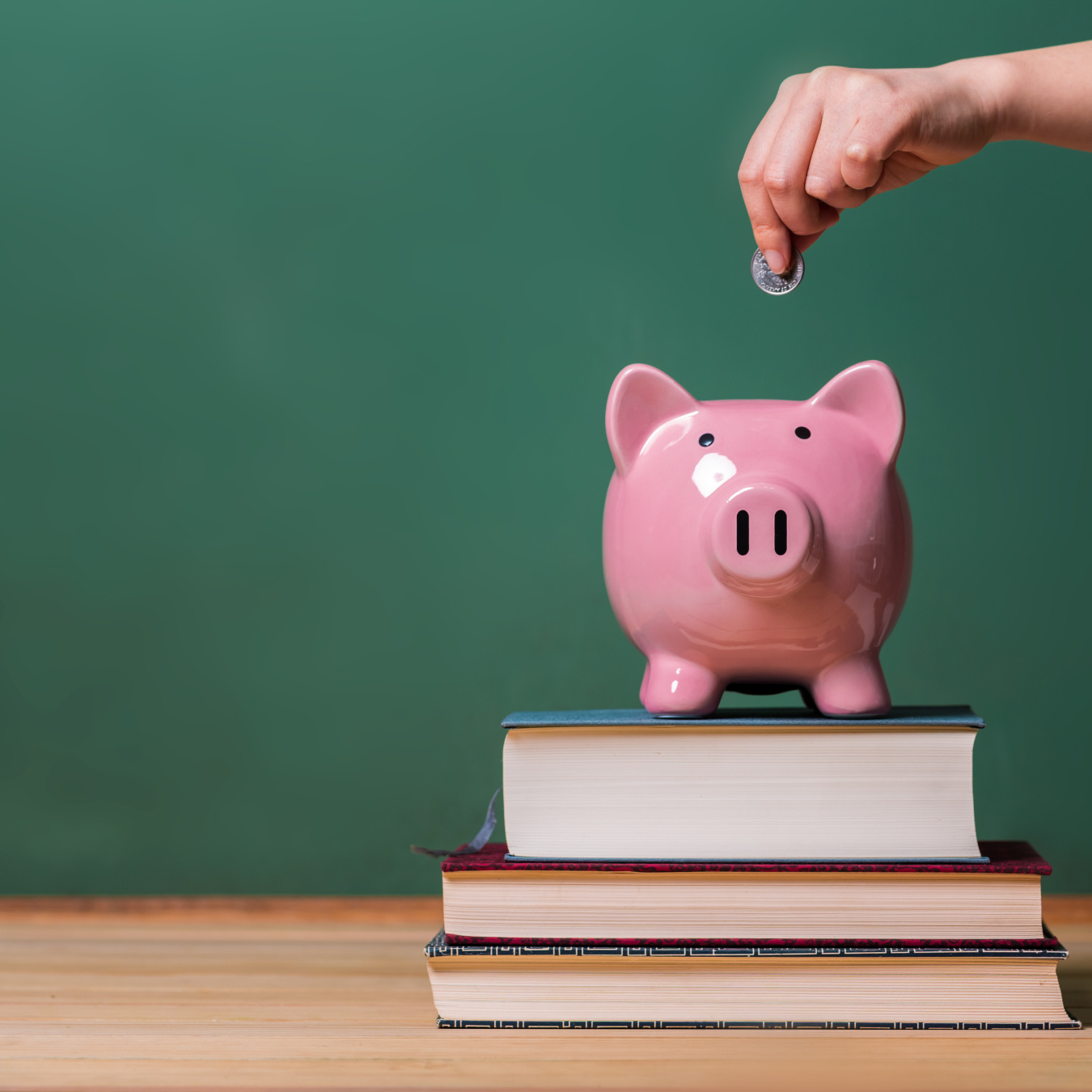 Piggy bank sitting on top of a small stack of books with a hand above it about to drop a coin in.