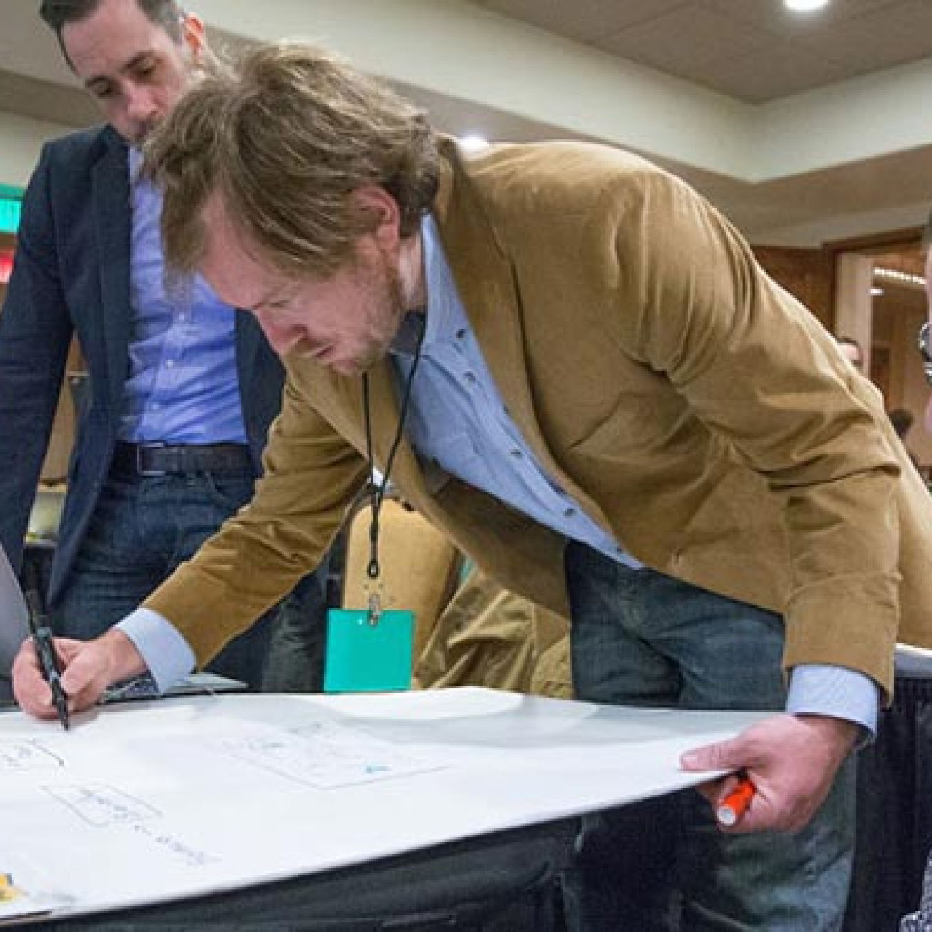 Image of three men working together at a table. The man in the middle writes on a large note pad while the other two men look on.