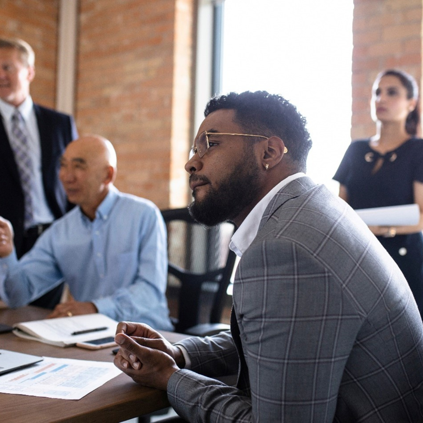 Four people of various ages sit and stand at a conference table looking out in the distance. 