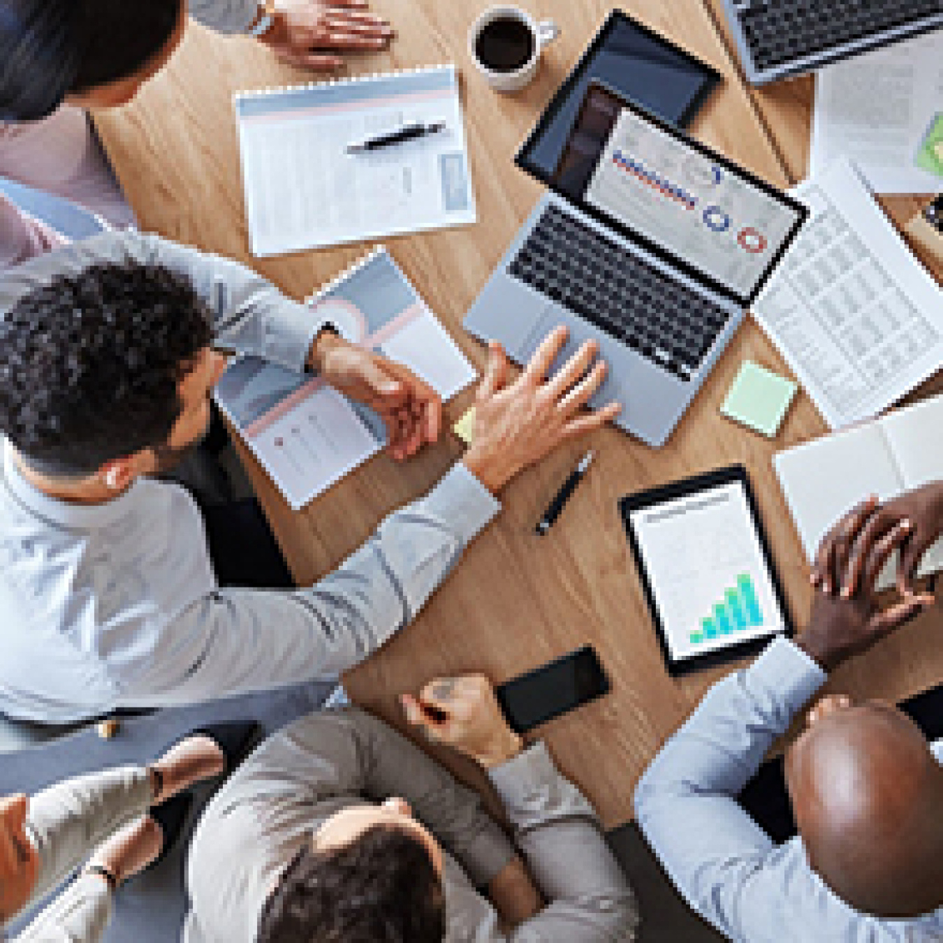 Photo looking down at a conference table surrounded by five people. On the table are laptops, tablets, and notebooks.