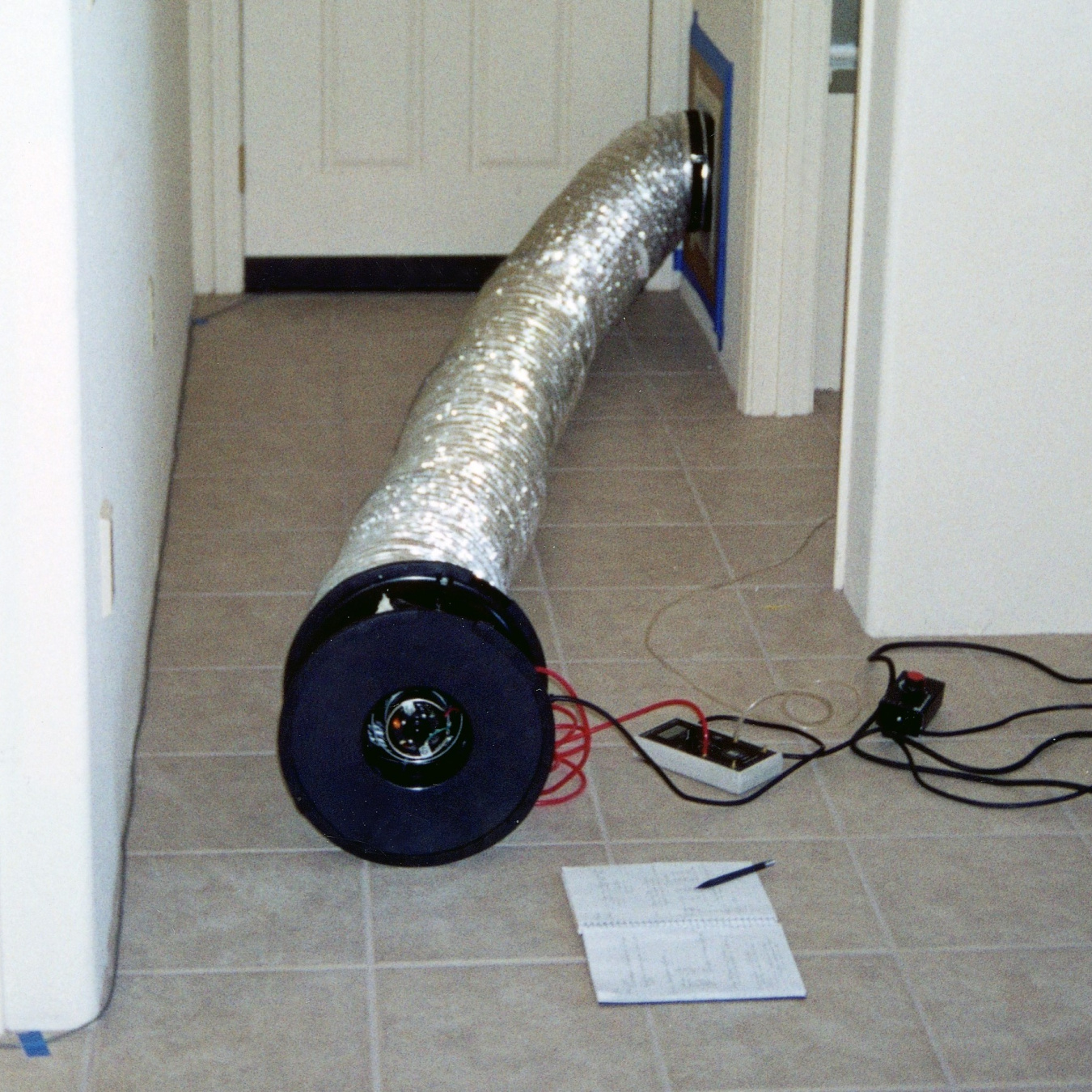 An air duct attached to cords and testing devices coming out of a wall in a hallway.
