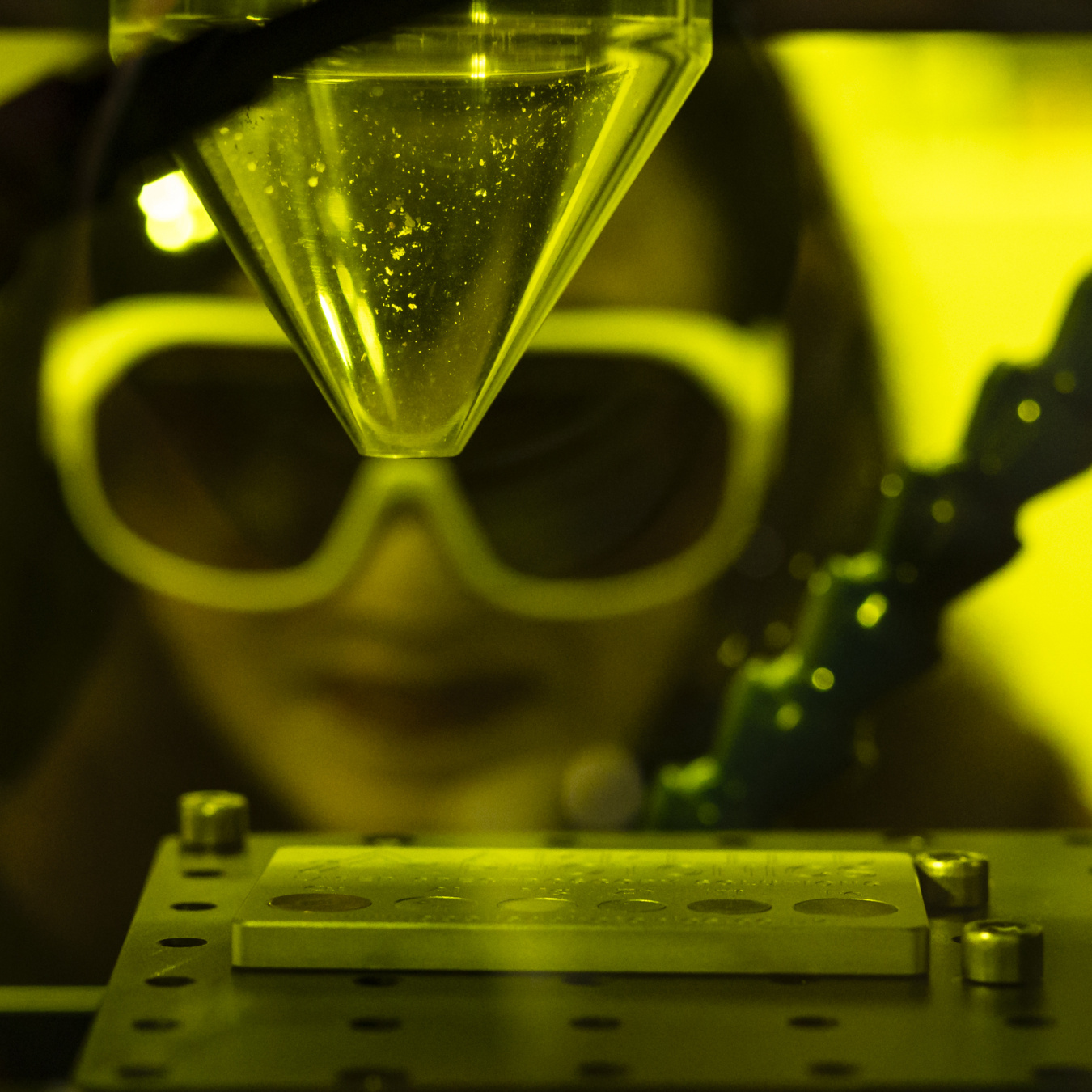 Madhavi Martin (a Southeast Asian woman with large lab goggles on) looks into a yellow-tinted box used for laser-induced breakdown spectroscopy. Inside the box is a clear funnel that is pointed towards a metal sheet with holes in it.