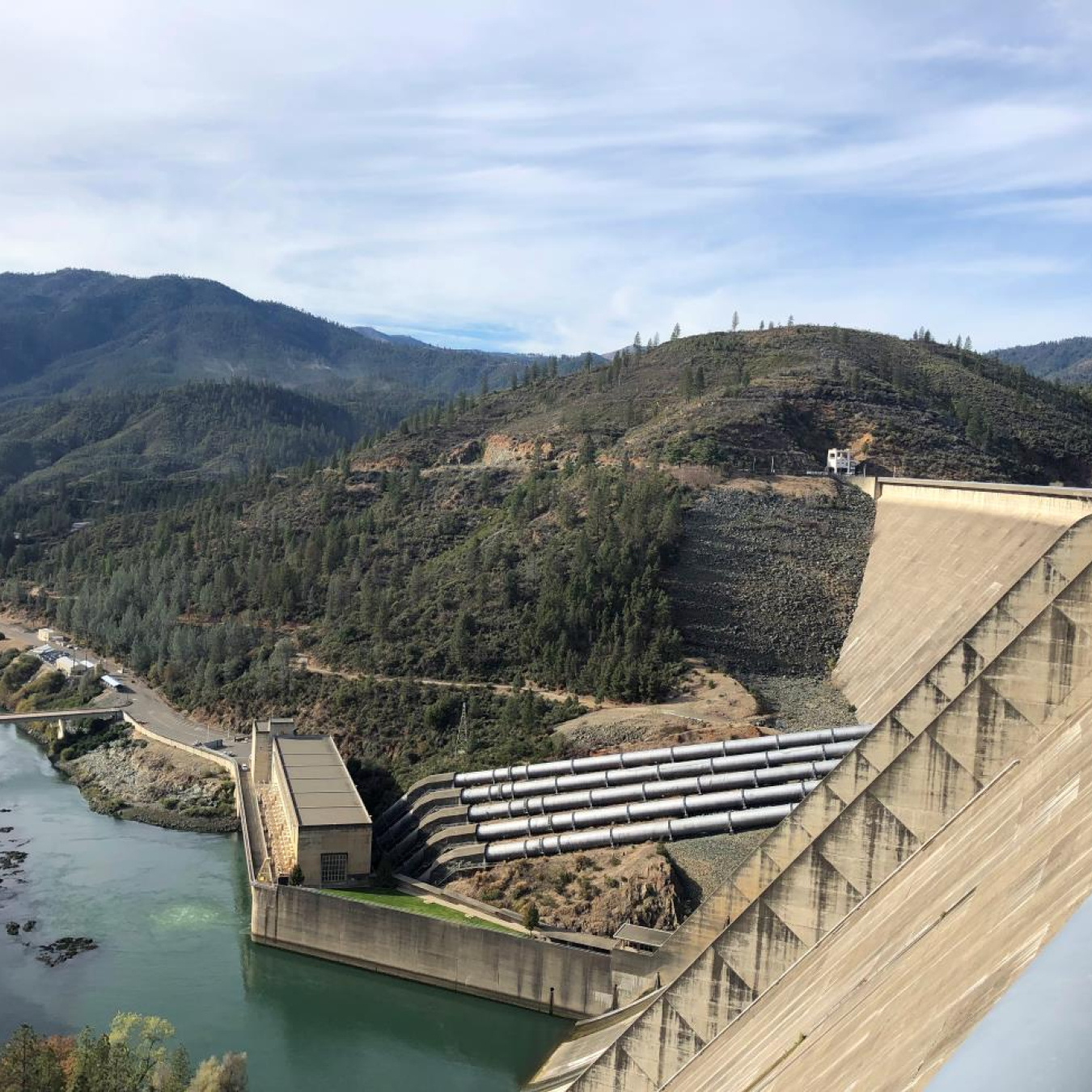 Side view of a large hydropower dam with the outflowing river and tree lined valley in the background