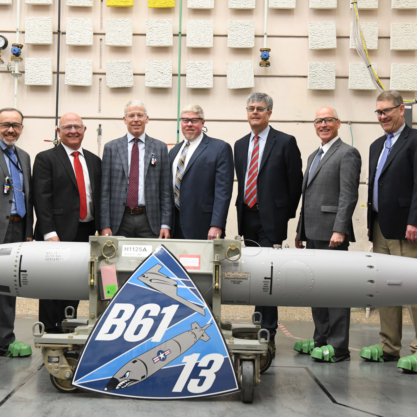 A group of men stand behind the first B61-13. In front of the bomb is an enlarged print of the B61-13 logo 
