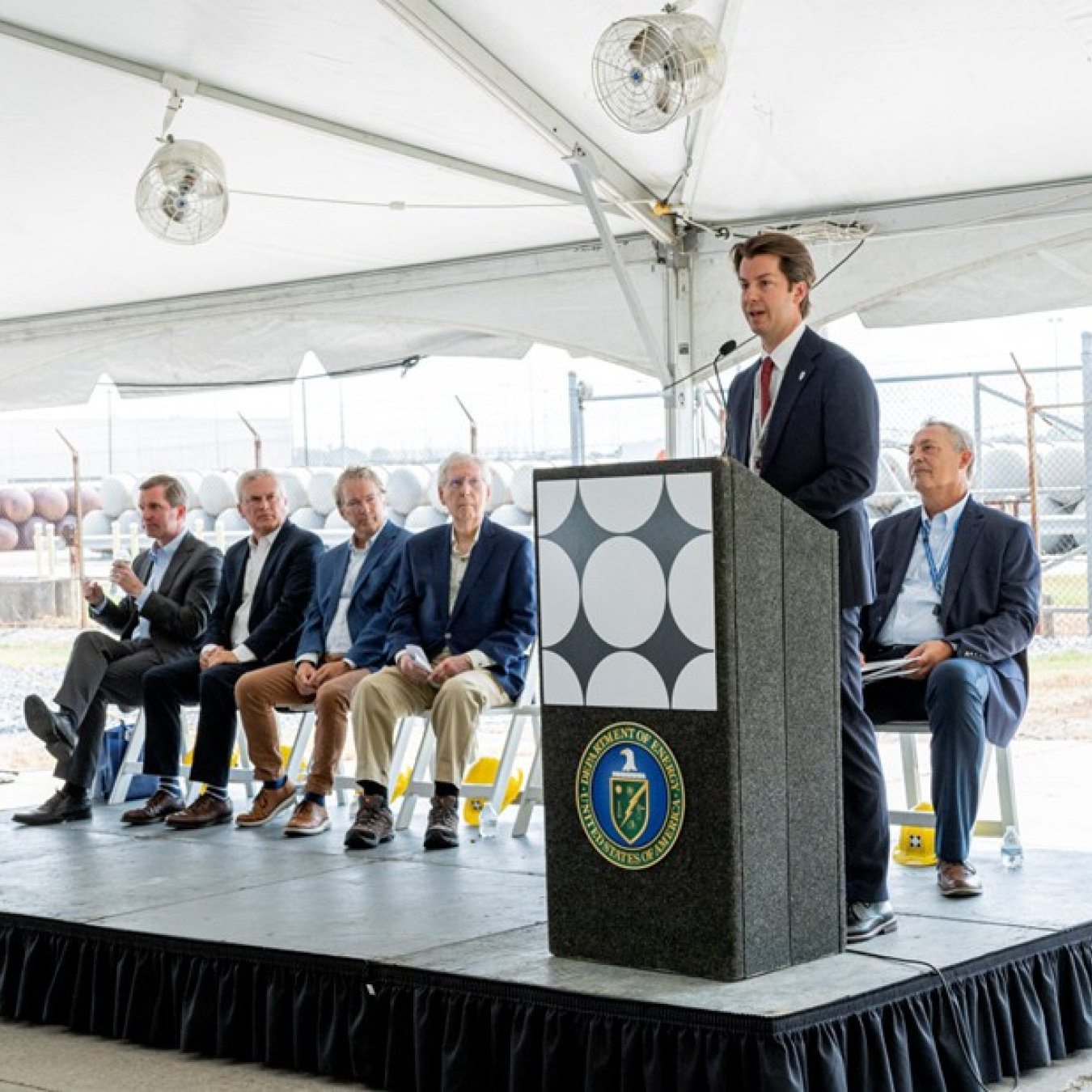 A man standing behind a podium giving a speech with five men sitting in chairs on the stage behind him