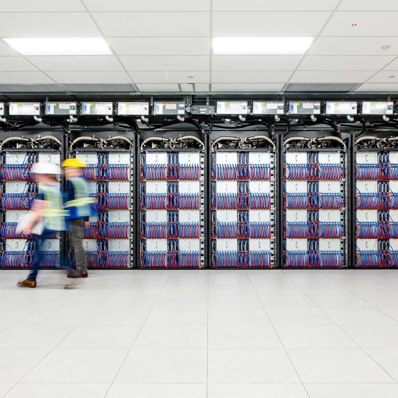A set of computer cabinets that are open with the red and blue tubes showing. Two people are walking past it and the photo was taken in a way that they appear blurred.