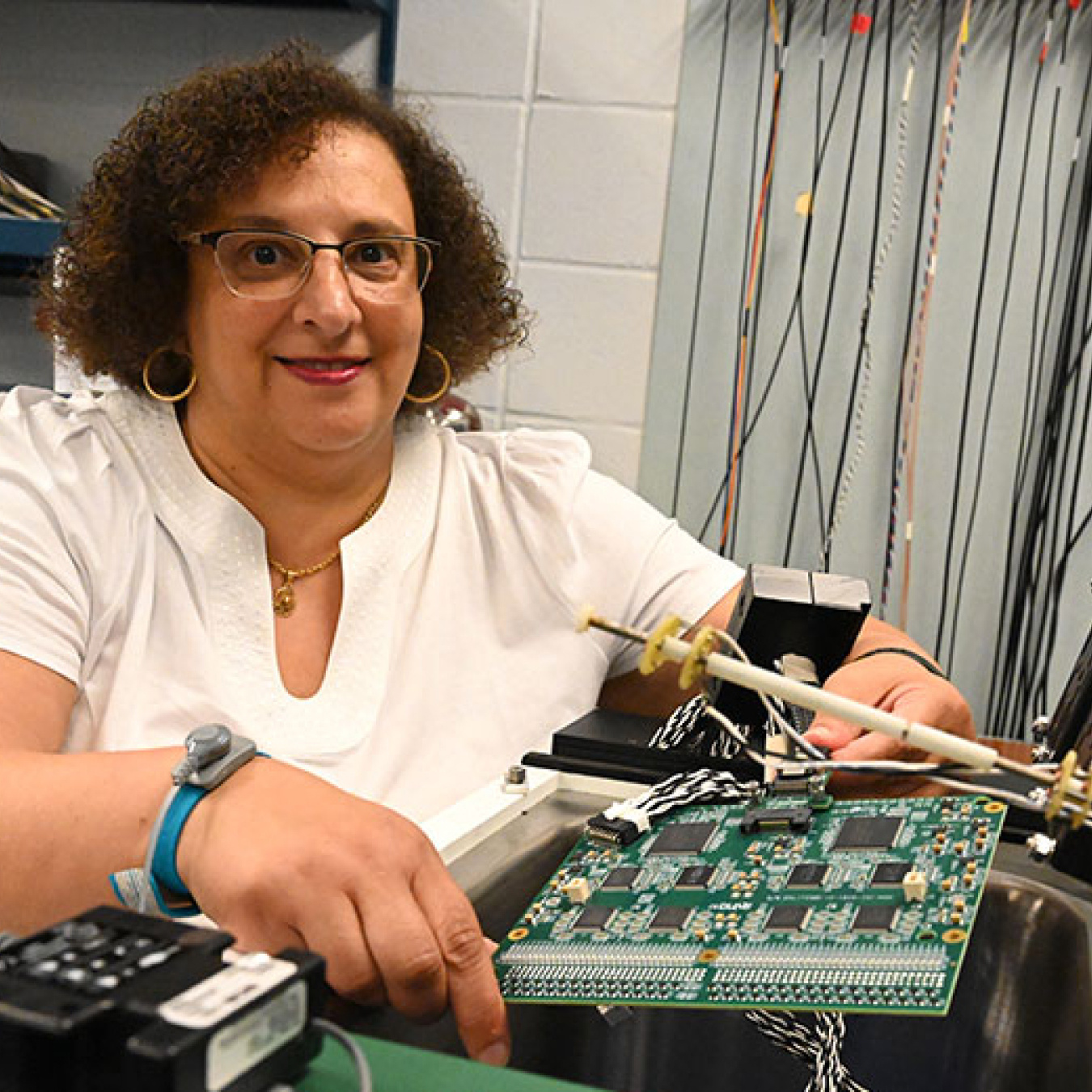 Mary Bishai (a woman in a white t-shirt) sitting at a desk with a microelectronics board in front of her and electronics equipment to the side.