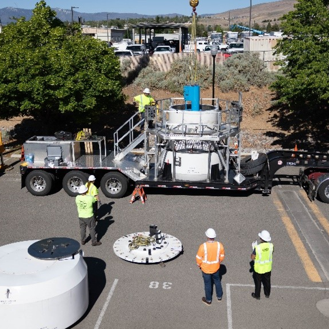 In a parking lot, a crane lowers a blue cylinder into a larger cylinder that is on a large trailer being towed by semi. Four people in high-visibility clothing and white hard hats watch.