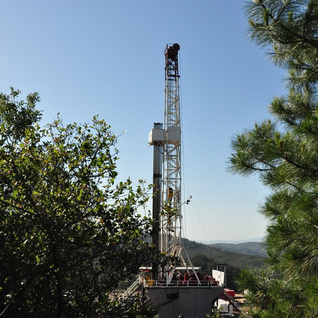 Photo of a drill rig between trees in the foreground, with rolling hills extending into the background.