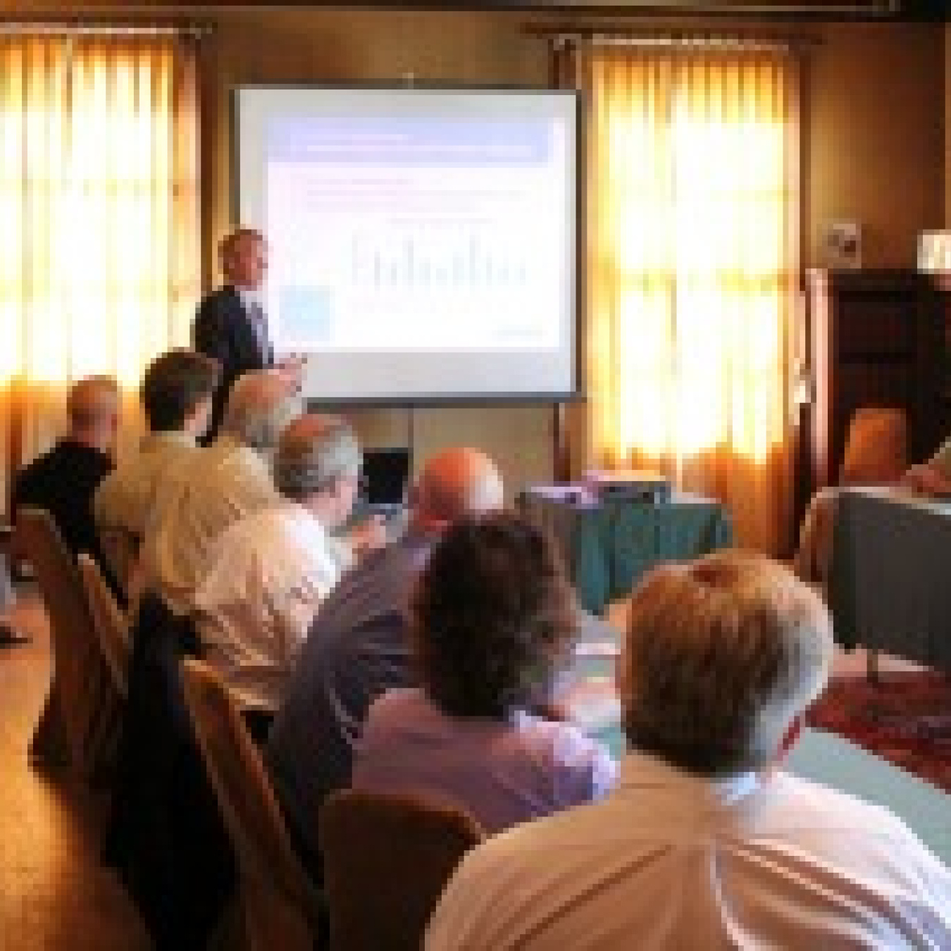 Group of people sitting at a table in a room, with a man speaking at the front and a display screen behind him.
