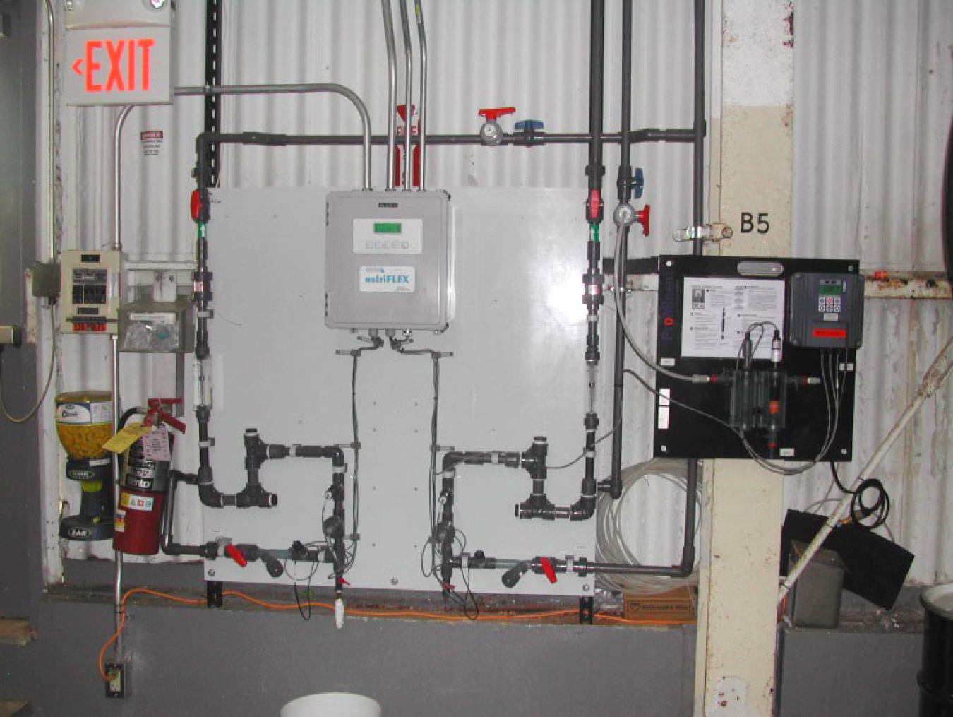 A metal wall has an advanced cooling tower control system mounted on it. There is a white control panel with series of pipes running in and out of it along the metal wall.