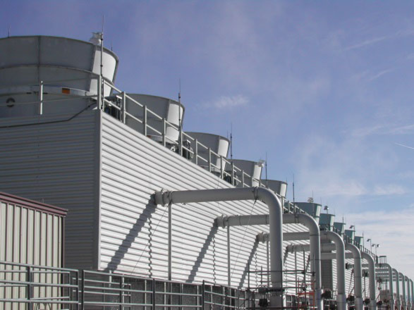 Image shows a silver metal building with a series of ten round gray cooling towers coming out of the roof. There is a blue sky in the background.