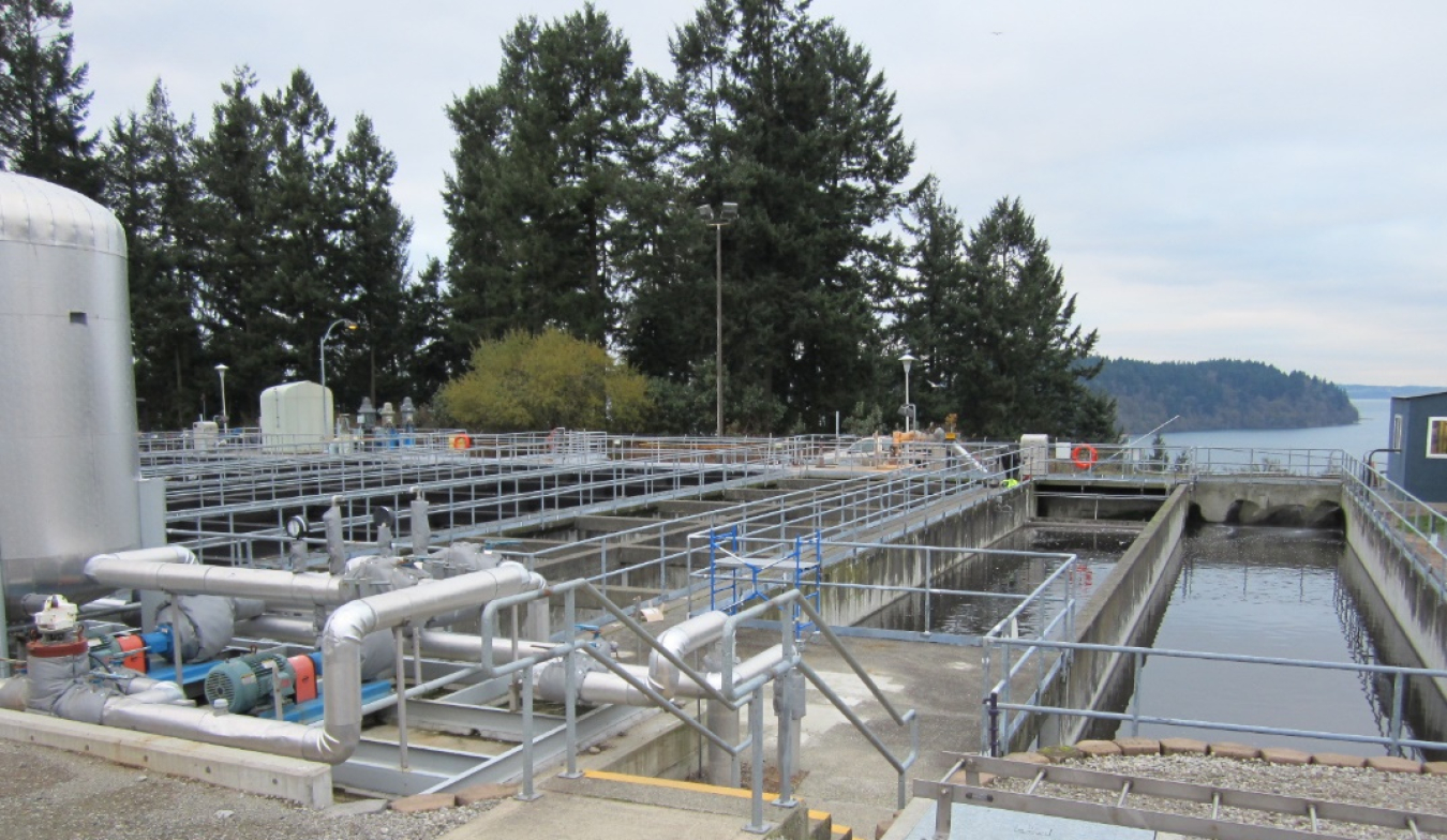 Image shows the tanks, pipes, and water holding areas of a wastewater treatment plan. Trees, shrubs, and a body of water are in the background.