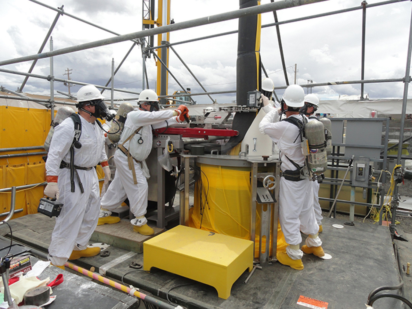 A worker, second from left, operates a mechanical lever to close foam insulation that wraps around the AW-106 pump column. At right, health physics technicians take readings prior to the operators installing the zip ties around the foam.