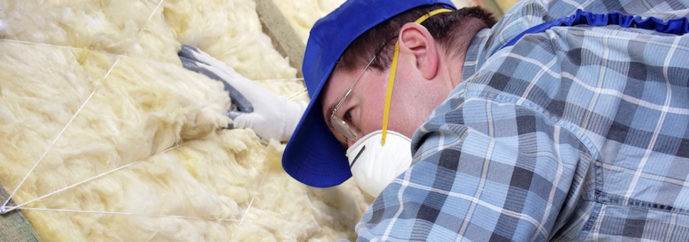 Photo of a man working on insulation in a building, wearing a hat and a face mask.