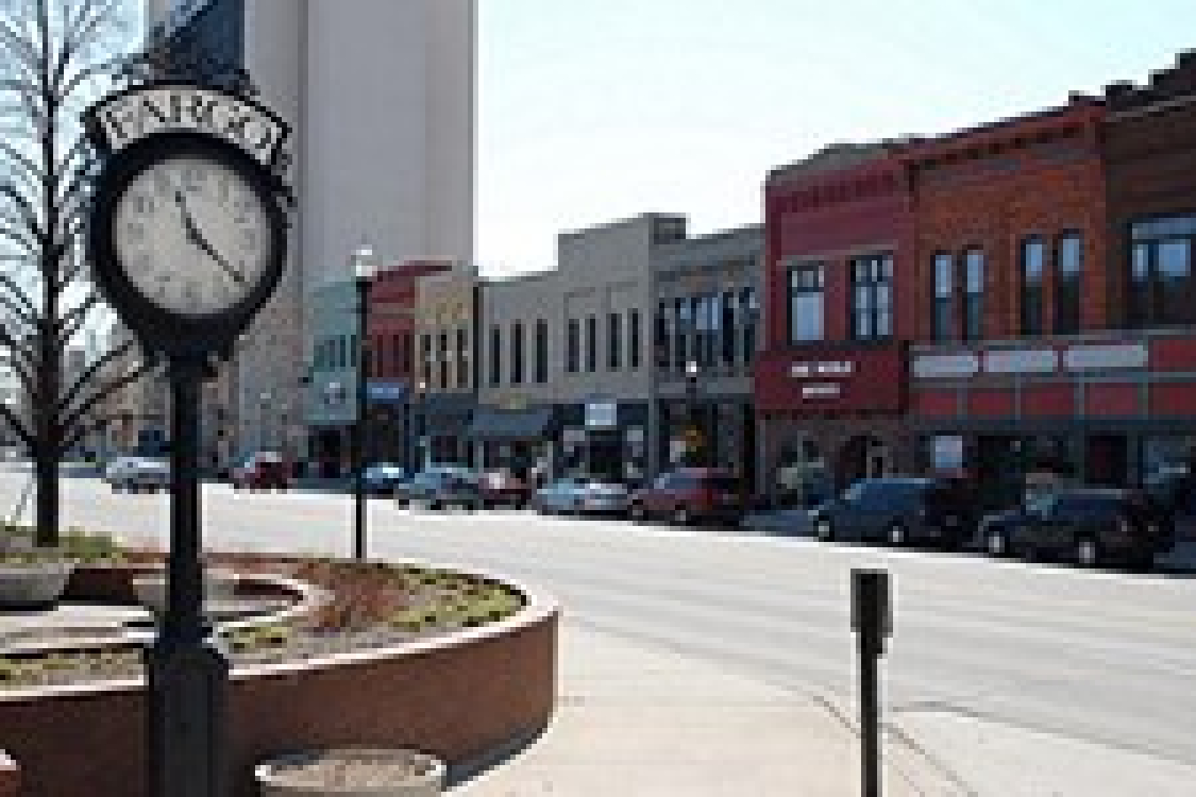 Photo of a city street, with a clock on a pole in the foreground and a block of businesses beyond.