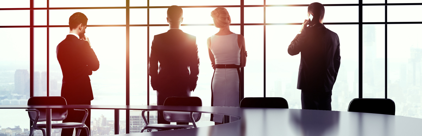 Photo of four people standing in front of a large window in an office's conference room or meeting room.