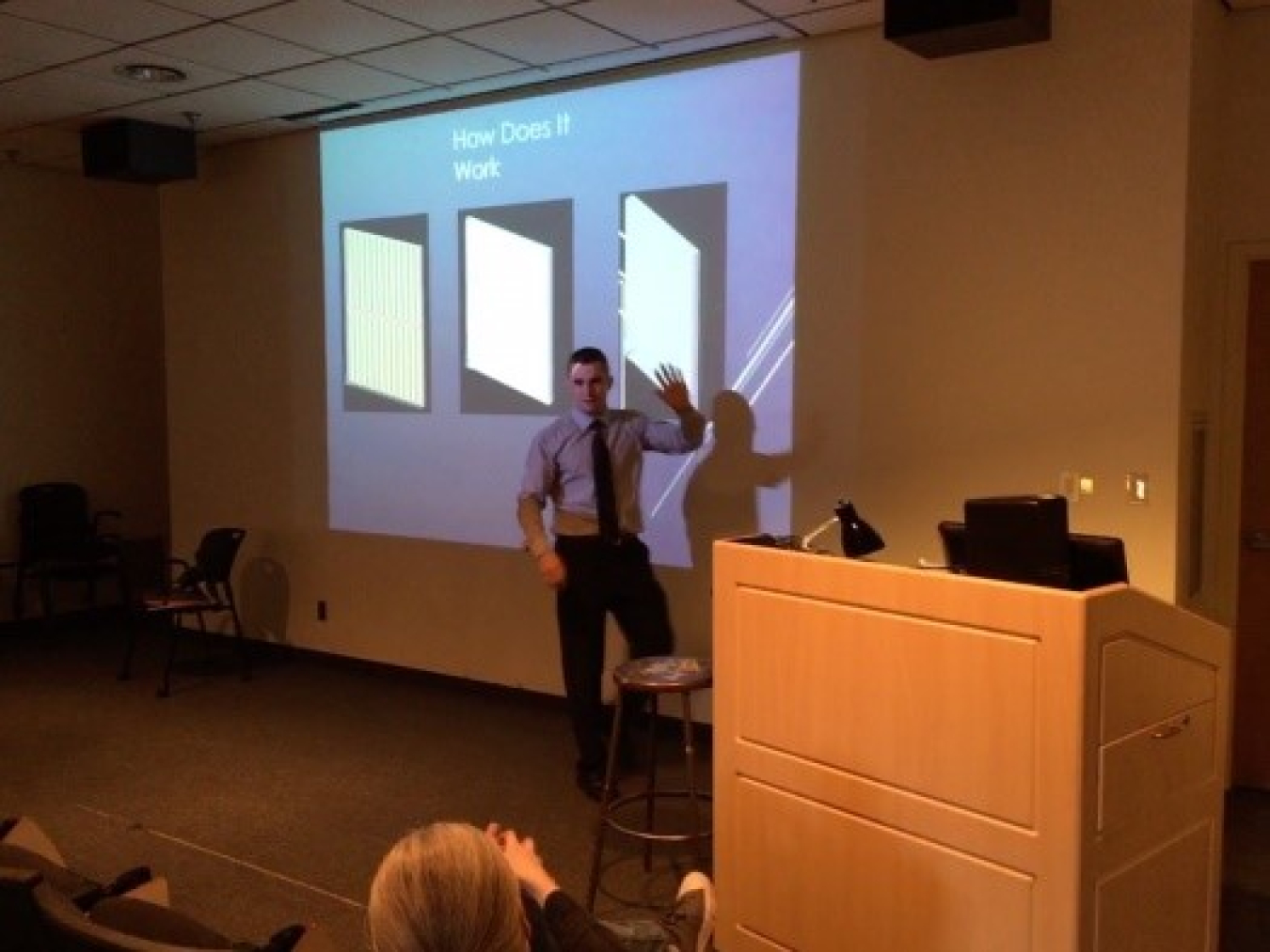 Photo of a man speaking at the head of a room near a podium, with a presentation on the large screen behind him.