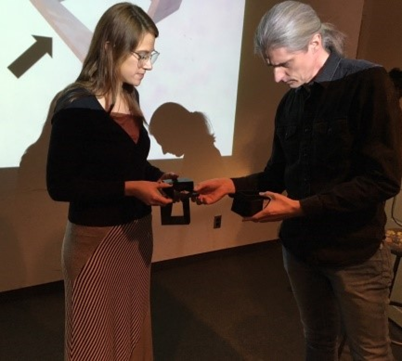 Photo of two people in a presentation room, with a screen behind them, examining objects for a project.