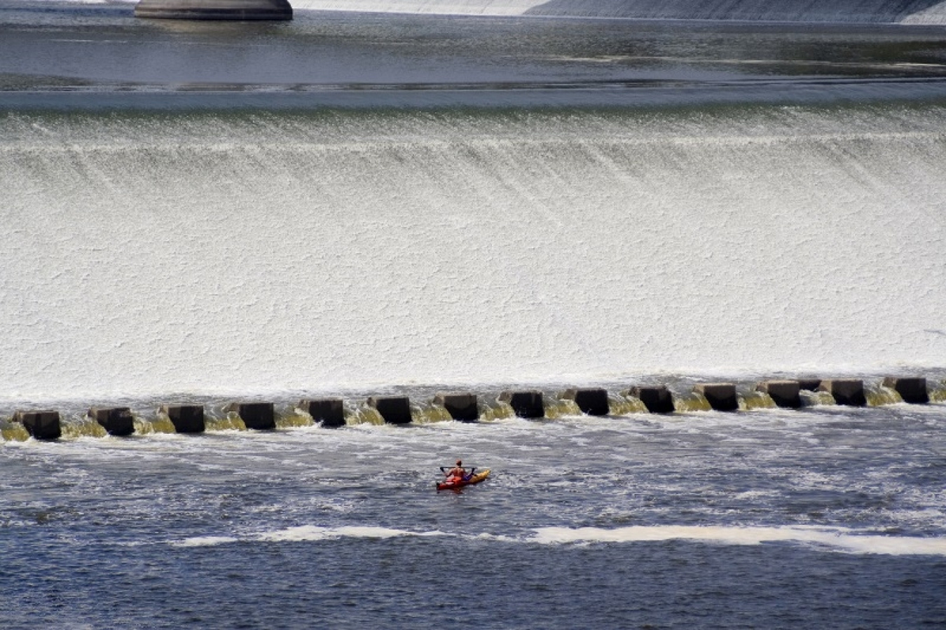 A kayaker paddles in the water in front of the water pouring off a dam.
