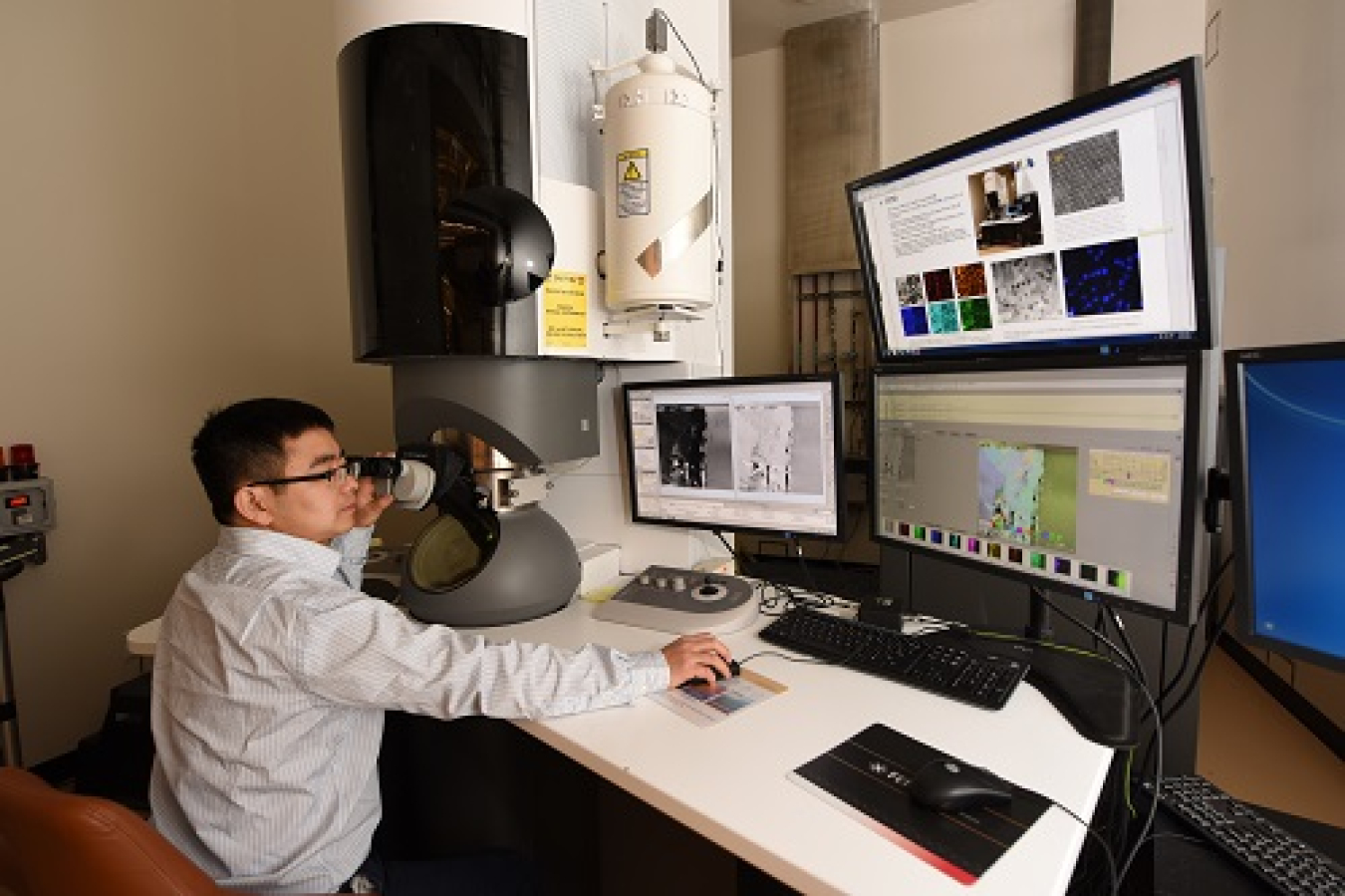 A scientist looking at computer screens with a microscope in the background.