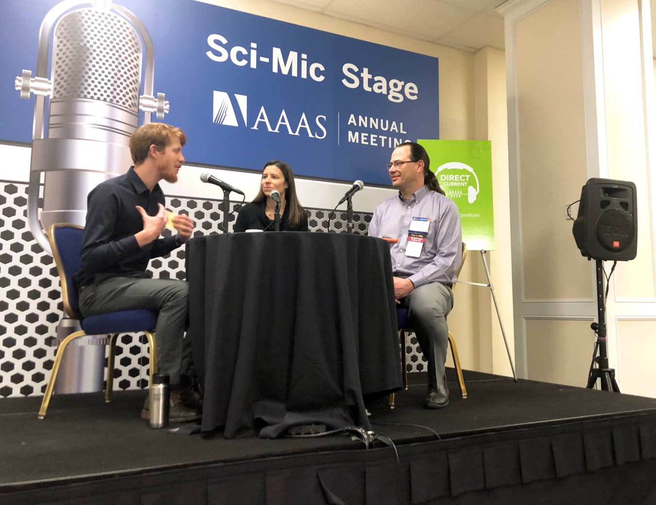 Three people sit around a table with microphones in front of a backdrop that reads "Sci Mic Stage AAAS Annual Meeting."