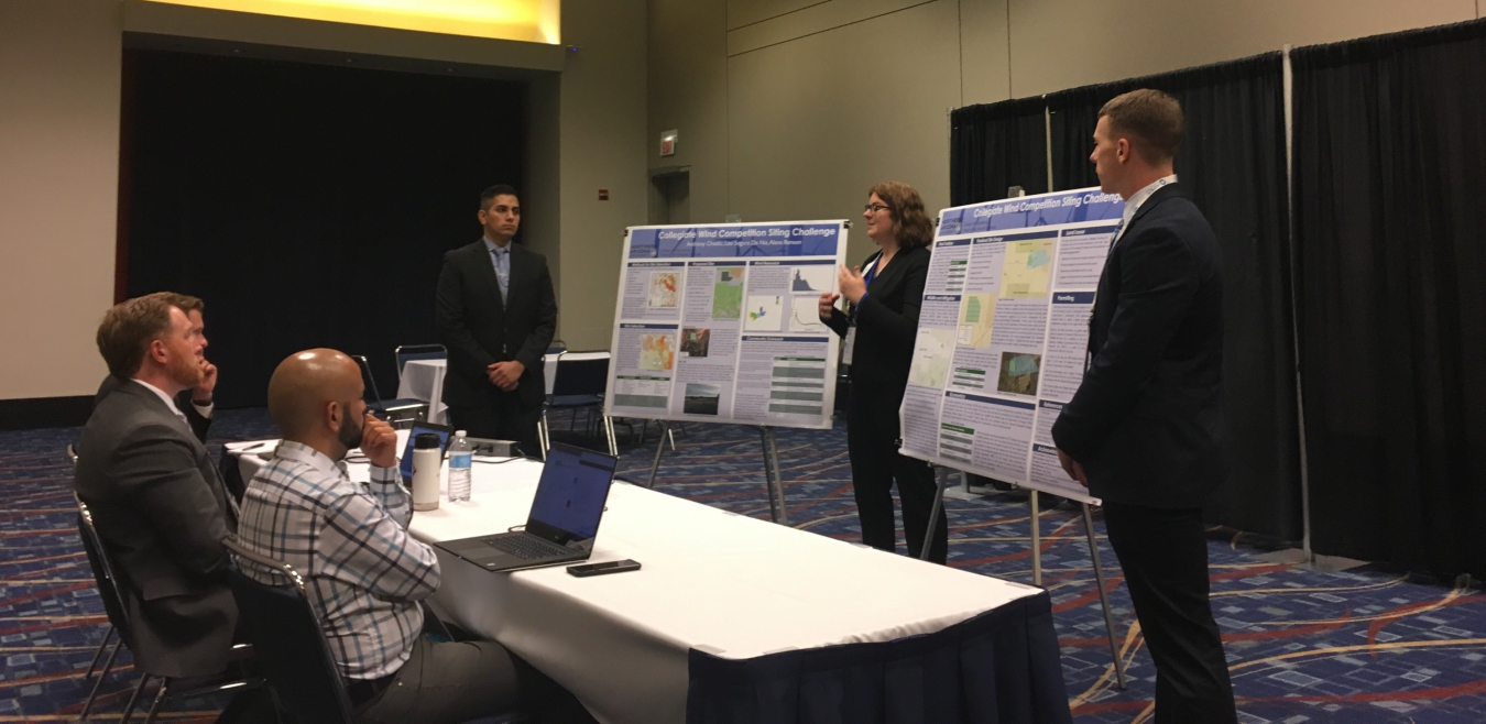 Three students stand next to two posters in front of a panel of judges. 