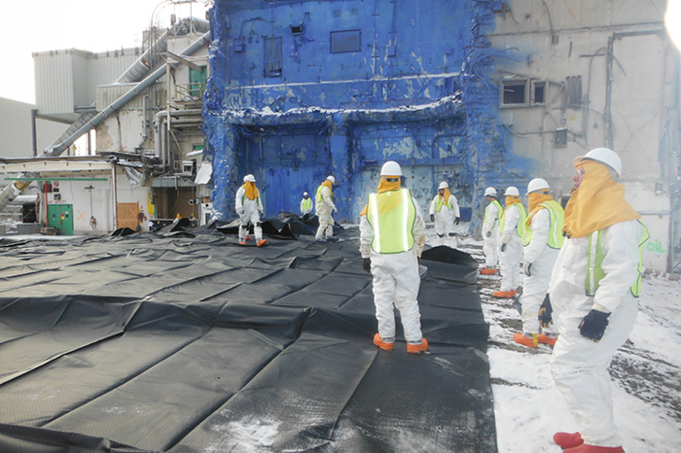 Workers pull and smooth out the cover over the Vitrification Facility concrete slab. The Main Plant, which is in the process of being deactivated, is in the background.
