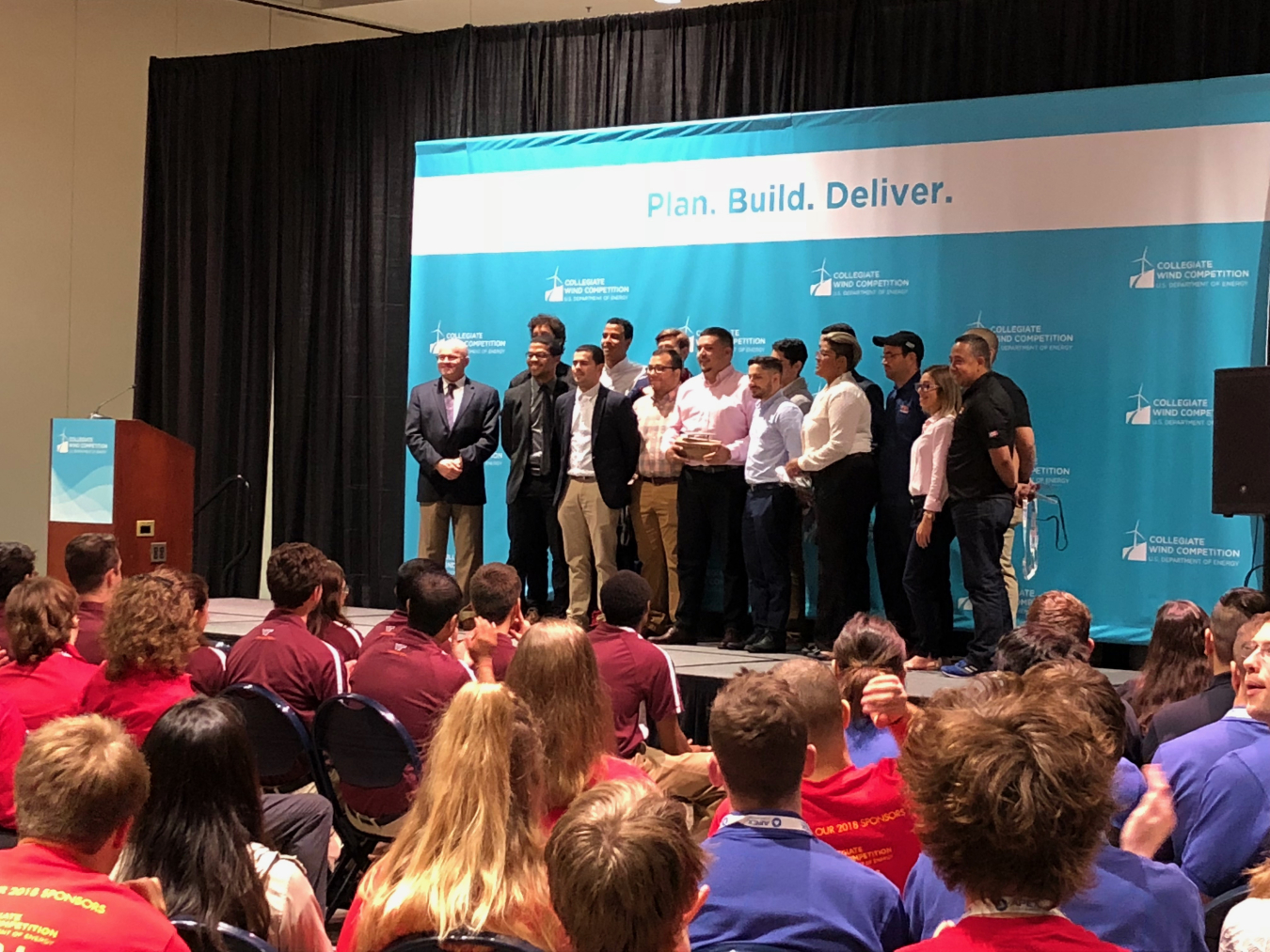 A student team in the Collegiate Wind Competition stands on stage in front of a banner at the WINDPOWER conference.