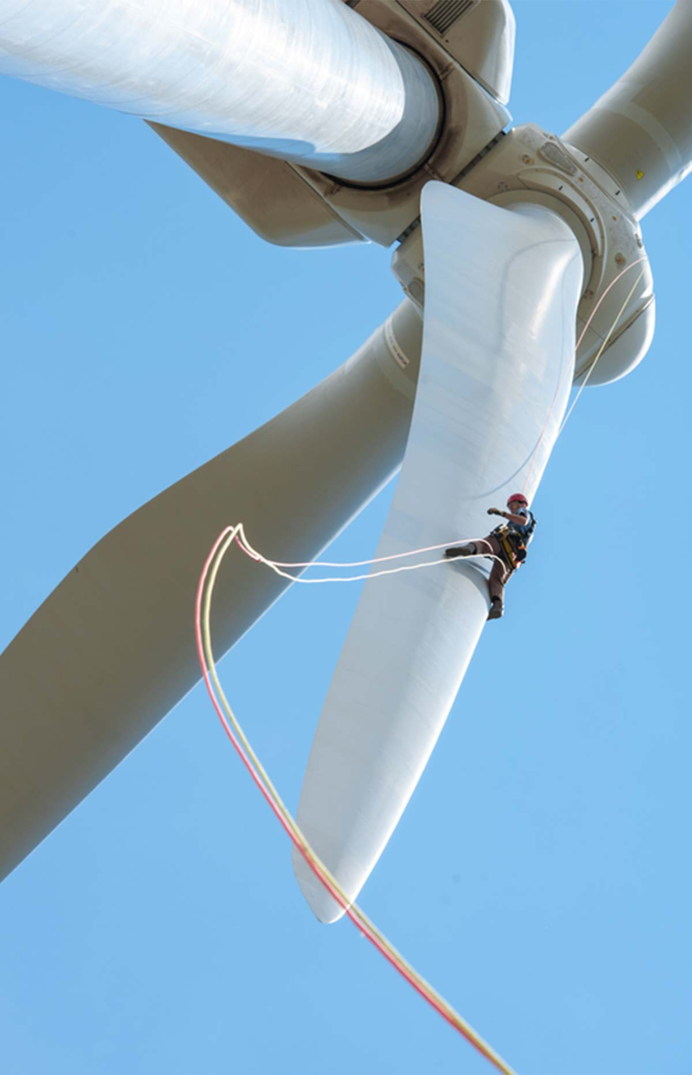 Large wind turbine blades and a man climbing one of the blades, with a rope dangling from beneath him.